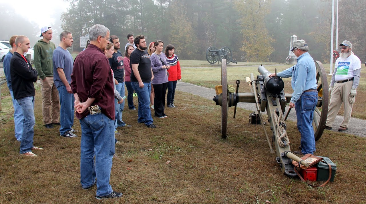 Members of the history department's Day of Remembrance group listen intently as Britton Lane Battlefield Association President Jim Weaver (second from the right) talks about the steps involved in firing a cannon. James Price of the battlefield association is on the right.