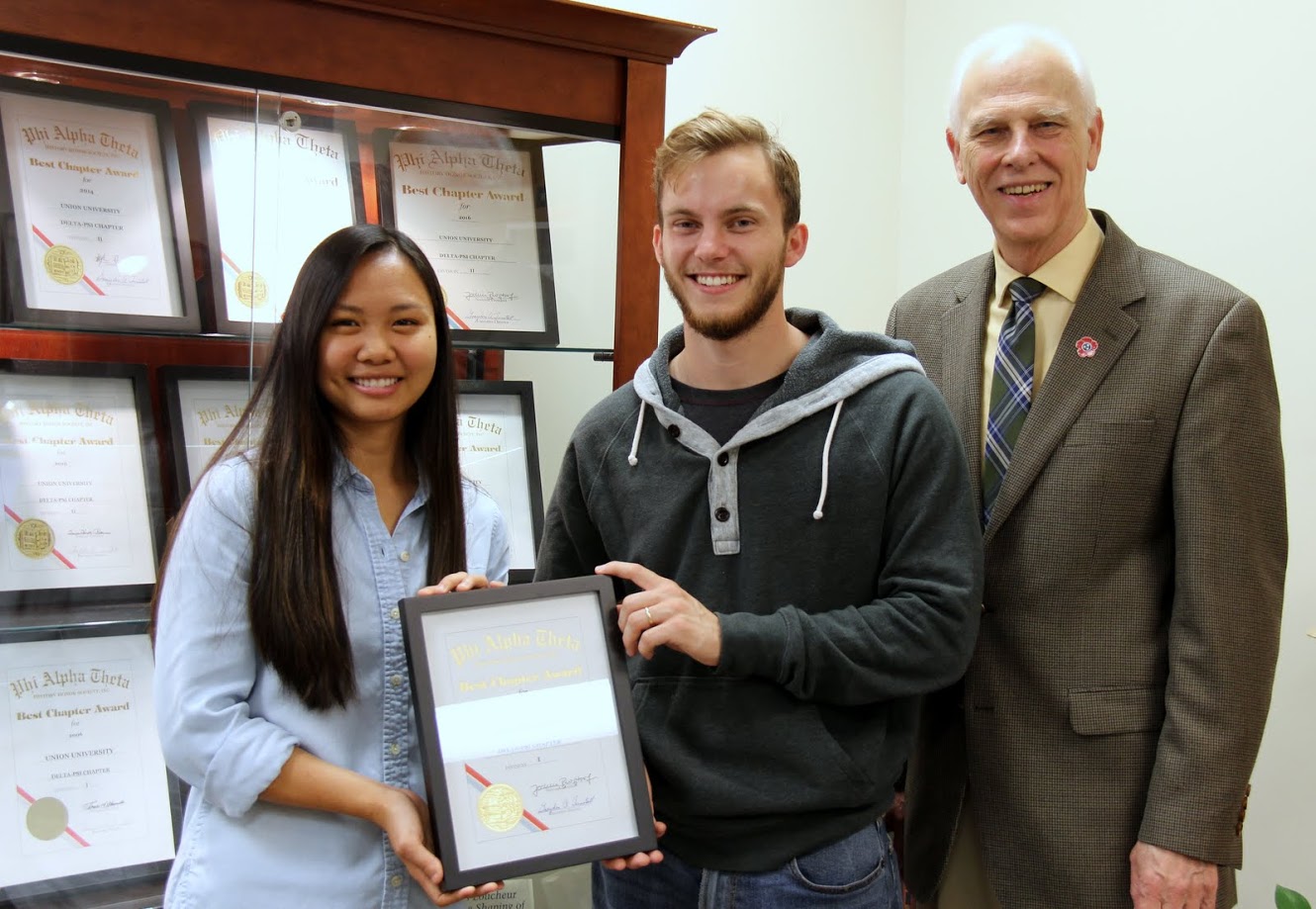 Delta-Psi chapter officers Katherine Cheshire and Seth Reid hold the 2017 Phi Alpha Theta Best Chapter Award certificate in front of the history department display case that contains other Delta-Psi Best Chapter Awards. Dr. Stephen Carls, the chapter faculty advisor, stands with them.