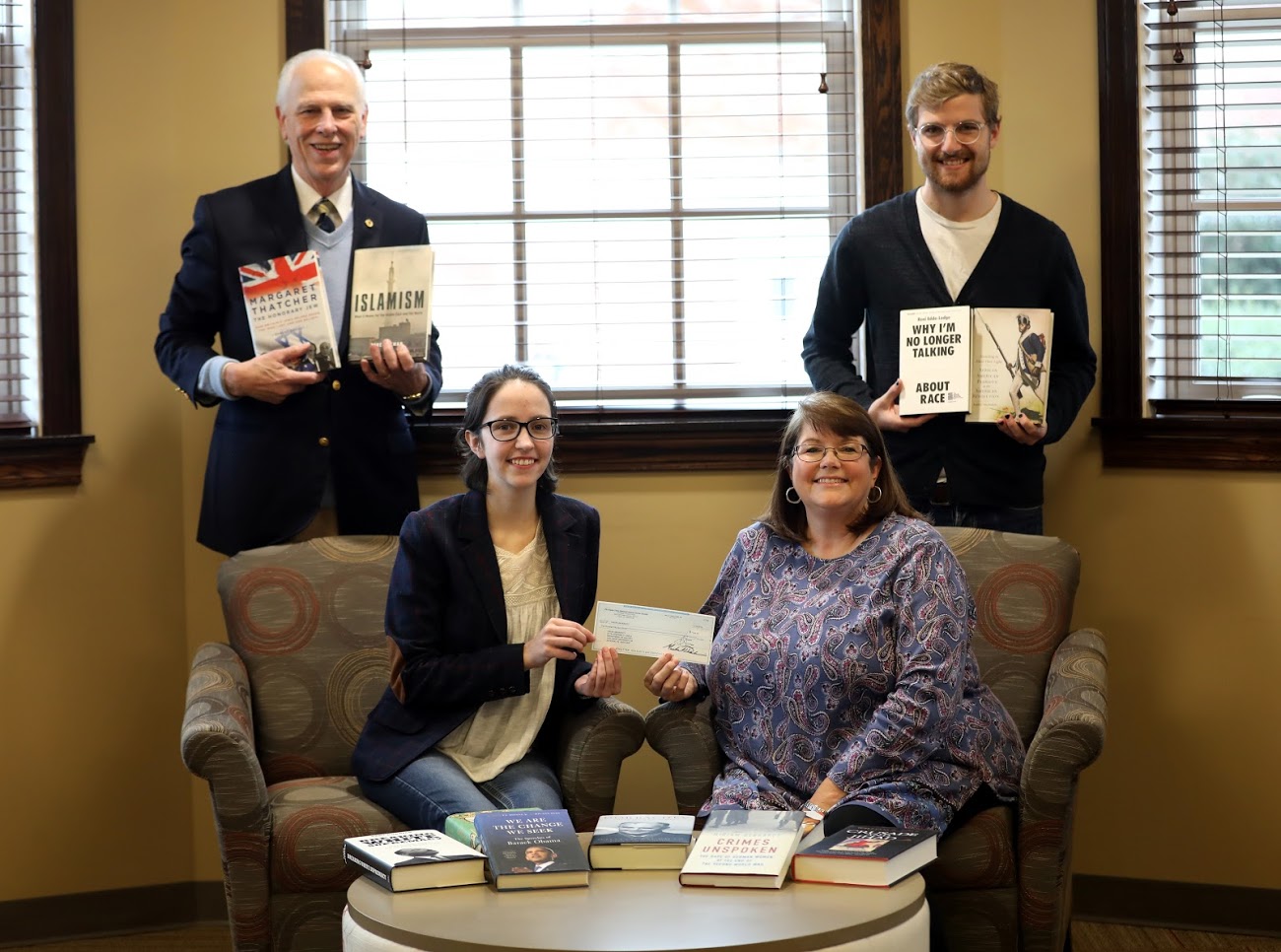 Delta-Psi Chapter President Lauren Butler (seated, left) presents the Phi Alpha Theta Best Chapter Award check to Library Director Melissa Moore on November 29, 2018. Faculty Sponsor Dr. Stephen Carls (standing, left) and Vice President Luke Sower hold some of the books that the chapter purchased with the prize money. Other purchased books are displayed on the front coffee table. Photo by Kristi Woody