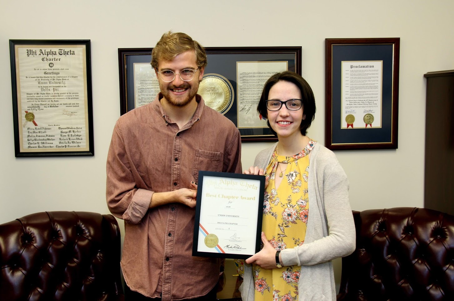 Delta-Psi Chapter Vice President Luke Sower and President Lauren Butler hold the 2018 Phi Alpha Theta Best Chapter Award that Delta-Psi received in the mail on October 18. Behind them are documents related to the chapter's history.