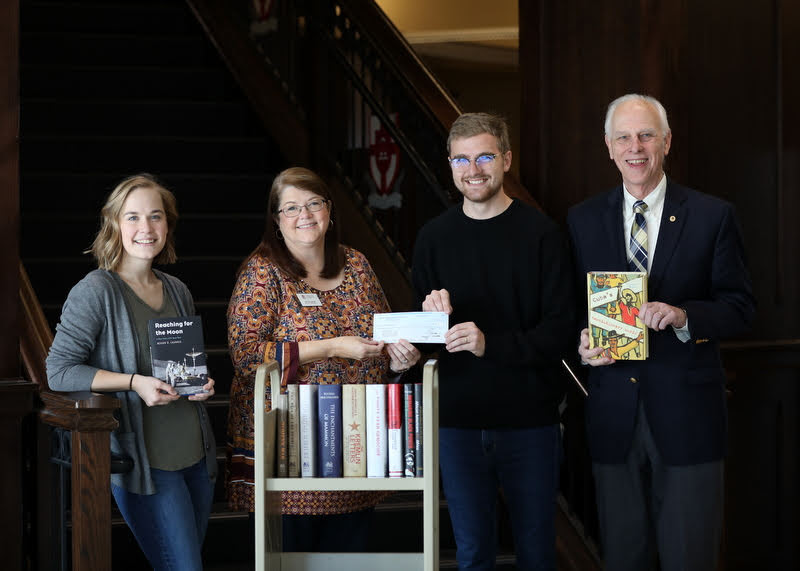 Delta-Psi Chapter President Luke Sower (second from right) presents the chapter's 2019 Phi Alpha Theta Best Chapter Award check of $250 to Library Director Melissa Moore (second from left) on December 5, 2019. Chapter Vice President Megan Ledbetter (left) and Dr. Stephen Carls (right) each hold one of the history books that the chapter purchased with the prize money. The other purchased books are on the book cart. Photo by Kristi Woody